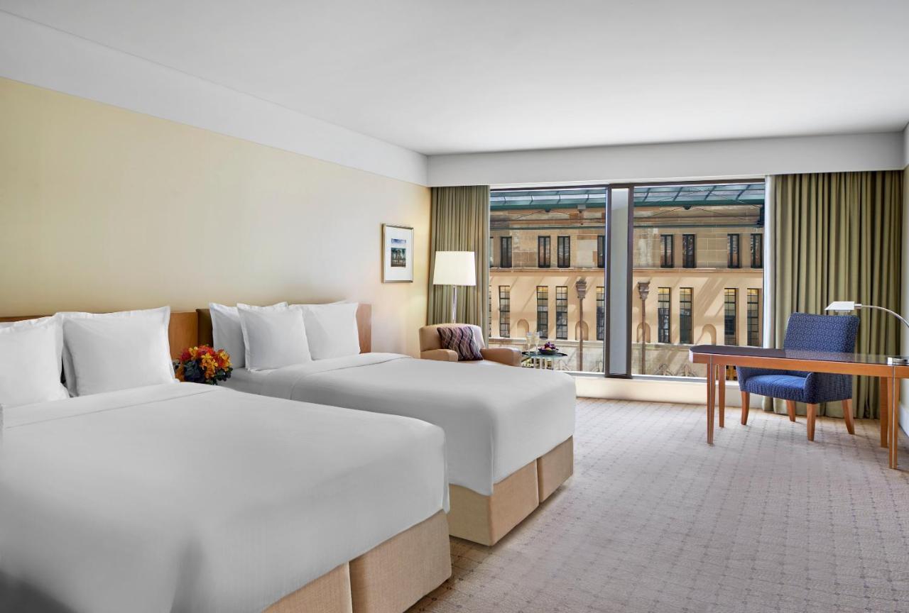 The Fullerton Hotel Sydney Esterno foto The photo shows a modern hotel room with two neatly made beds, each adorned with white bedding and pillows. There is a bedside table with a small decorative arrangement and a lamp beside one of the beds. The room features large windows with sheer cur