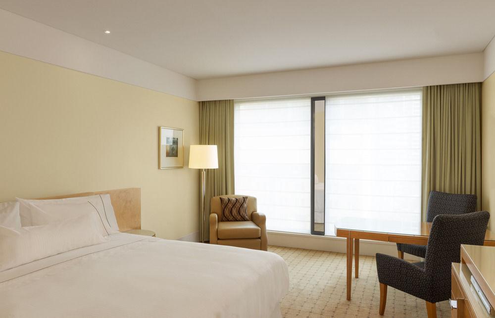 The Fullerton Hotel Sydney Esterno foto The photo shows a modern hotel room. The decor features soft, neutral colors, creating a calm and inviting atmosphere. There is a large bed with crisp white linens against one wall. A bedside lamp stands on a small table next to the bed. 

Near the w