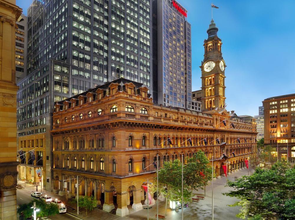 The Fullerton Hotel Sydney Esterno foto The image depicts a contrasting architectural scene featuring a historic building alongside modern skyscrapers. The foreground shows a grand, ornate structure with decorative elements, likely a former post office or civic building, distinguished by i