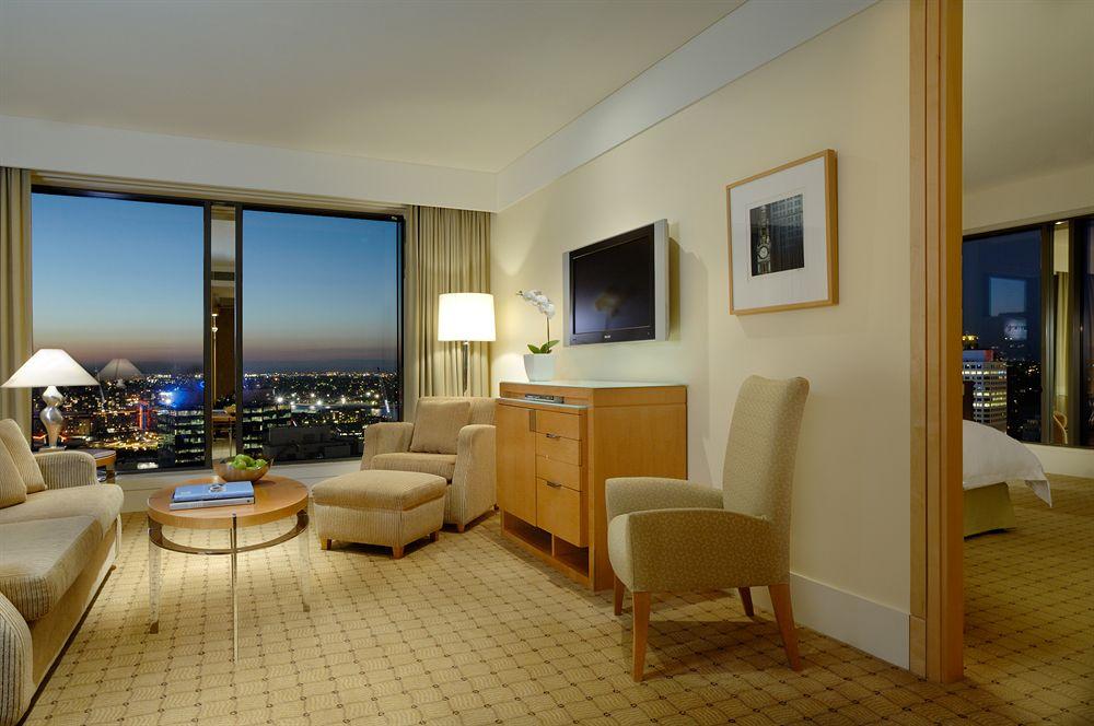 The Fullerton Hotel Sydney Esterno foto The photo shows a hotel room with a modern and elegant design. There's a large window with a view of a city skyline, indicating it's likely at a higher floor. The room features a seating area with two light-colored chairs and a small round coffee tab