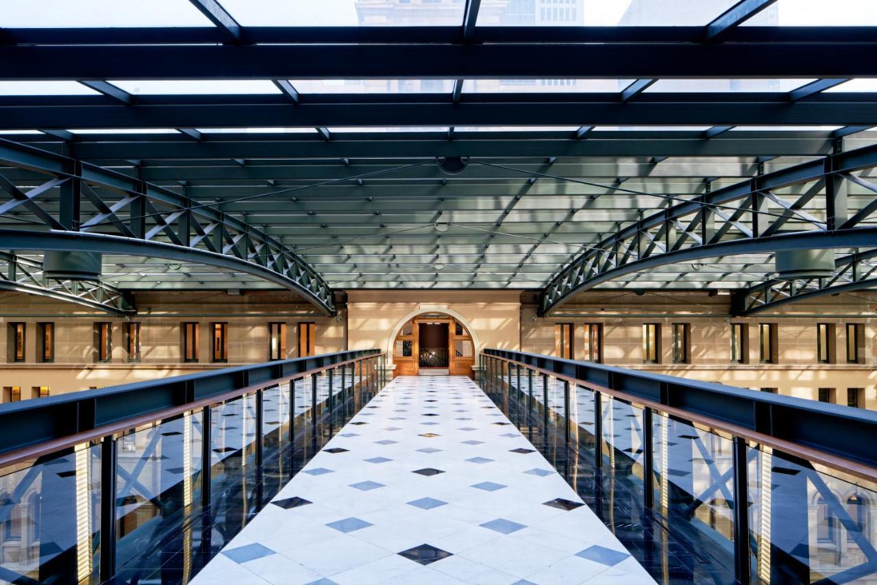 The Fullerton Hotel Sydney Esterno foto The photo shows an interior space featuring a modern architectural design. There is a wide corridor or walkway made of patterned tiles, with a series of geometric shapes visible on the floor. Above, a glass ceiling allows natural light to flood the a
