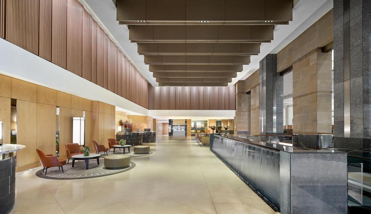 The Fullerton Hotel Sydney Esterno foto The photo shows a spacious and modern hotel lobby. It features high ceilings with large, rectangular light fixtures hanging above. The walls are a combination of warm wood tones and stone, creating an elegant atmosphere. On one side, there are comfor