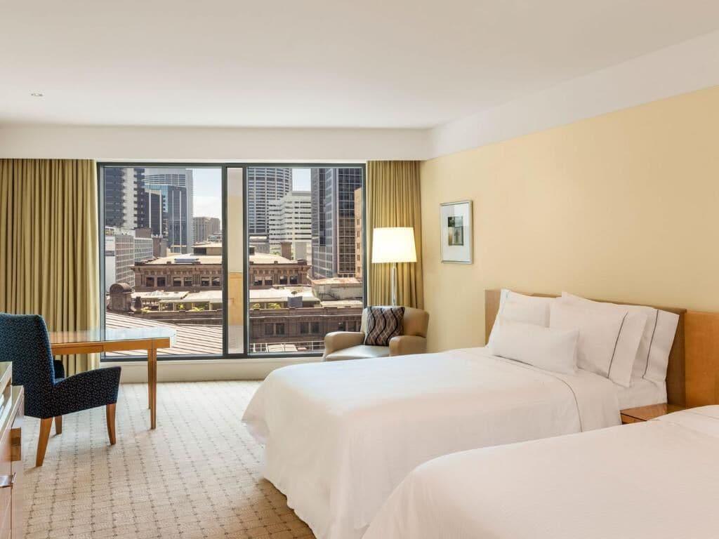 The Fullerton Hotel Sydney Esterno foto The photo shows a hotel room with a modern design. There are two beds with white linens positioned in the center. A large window offers a view of a cityscape, featuring tall buildings outside. To the side, there is a comfortable armchair and a wooden