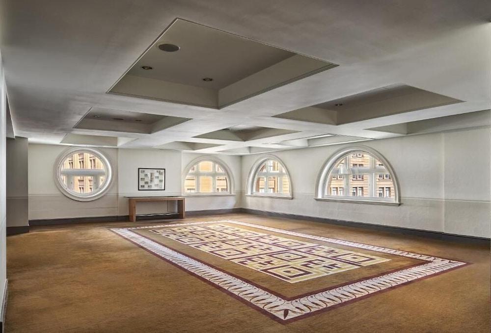 The Fullerton Hotel Sydney Esterno foto The photo shows a spacious, well-lit room with a contemporary design. The ceiling is divided into sections with recessed lighting. There are several rounded windows that allow natural light to enter, creating an airy atmosphere. A large area rug with