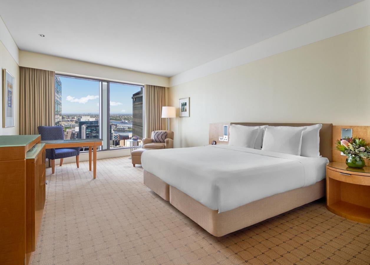 The Fullerton Hotel Sydney Esterno foto The photo shows a hotel room featuring a large bed with white linens, set in a spacious area with light-colored walls. To one side, there is a comfortable chair and a small table, both positioned near a large window that allows for natural light and 