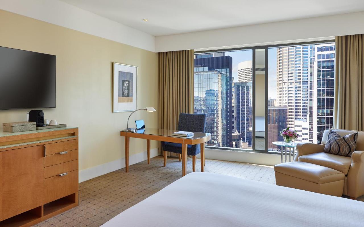 The Fullerton Hotel Sydney Esterno foto The photo shows a modern hotel room with a cozy and contemporary design. There is a comfortable bed with white linens, a desk with a chair and a lamp positioned by a large window that offers a view of a cityscape filled with tall buildings. A flat-sc