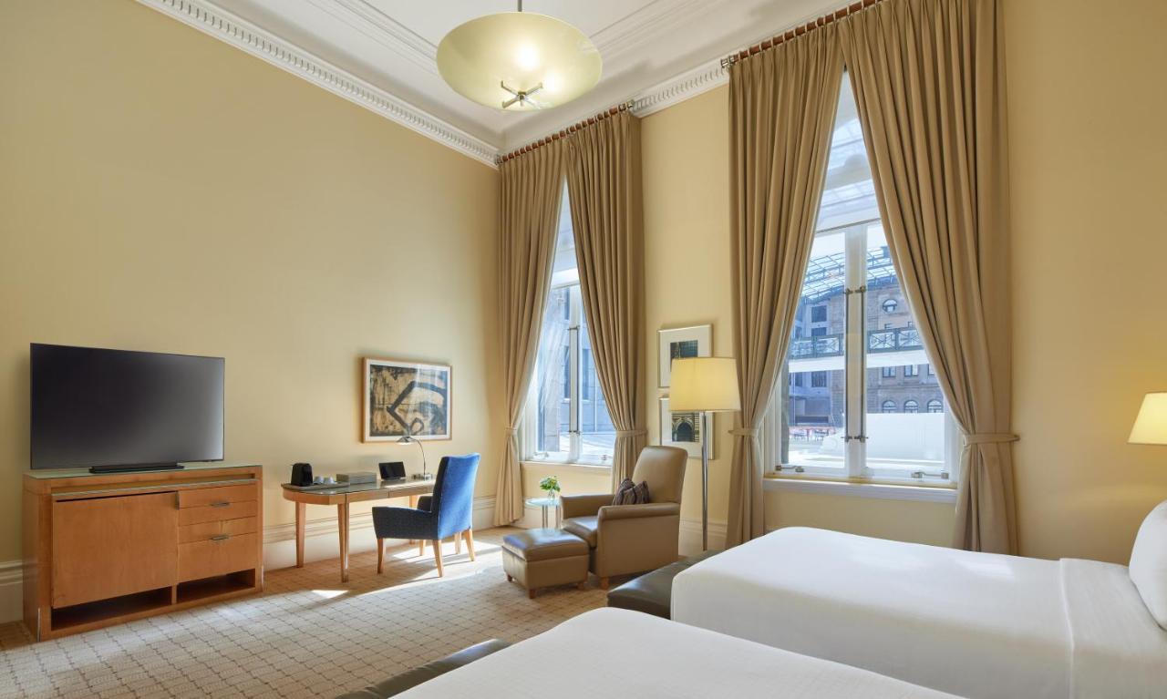 The Fullerton Hotel Sydney Esterno foto The photo shows a hotel room featuring a light-colored interior with high ceilings. There are two beds positioned at the forefront of the image, with a small bedside table between them. To one side, a wooden dresser supports a flat-screen television.