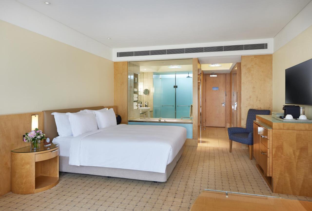 The Fullerton Hotel Sydney Esterno foto The photo shows a modern hotel room featuring a spacious layout. It includes a large bed with white linens, a wooden bedside table with a flower arrangement, and a chair in the corner. The room has a light-colored, neutral palette, giving it a clean 