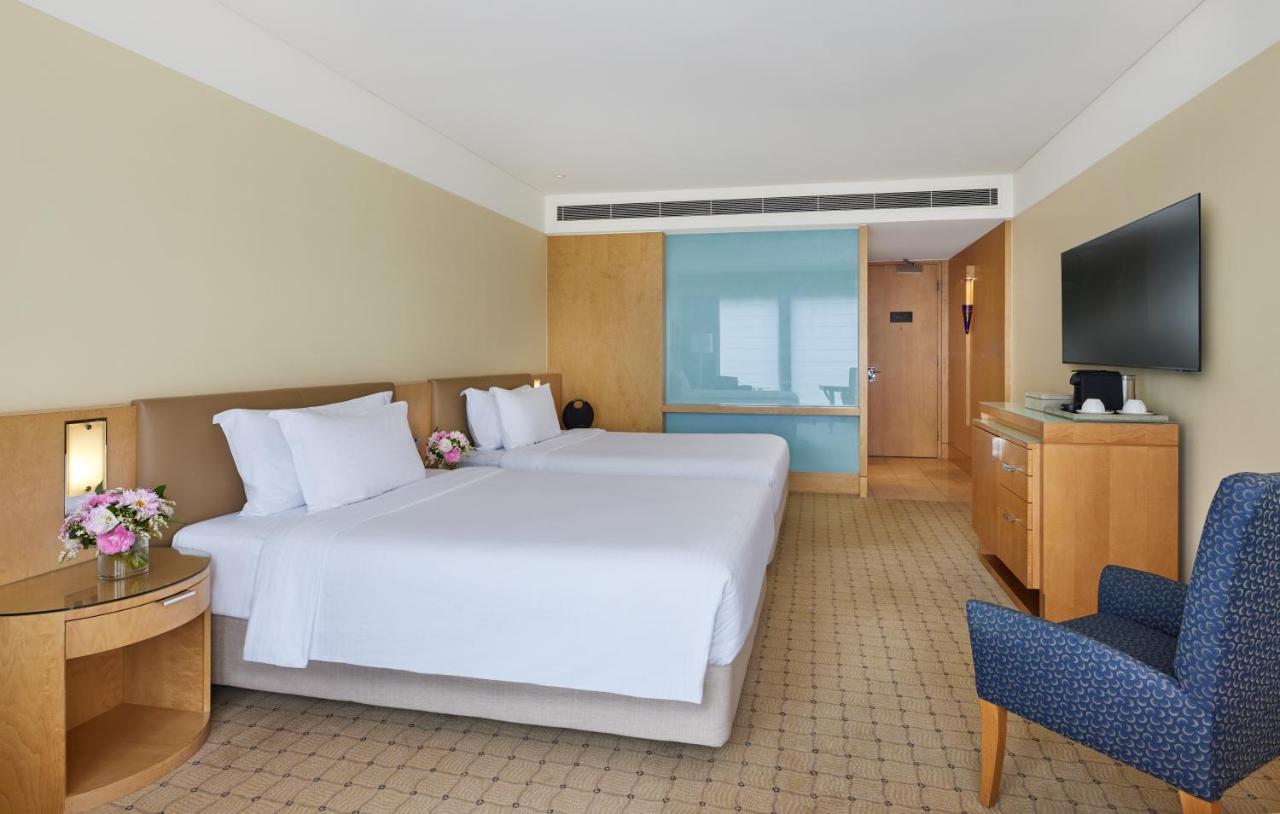 The Fullerton Hotel Sydney Esterno foto The image shows a hotel room featuring two twin beds with white linens and decorative pillows. The walls are painted in a soft beige color, and there is wooden furniture, including a bedside table and a media console. A flat-screen TV is mounted on t
