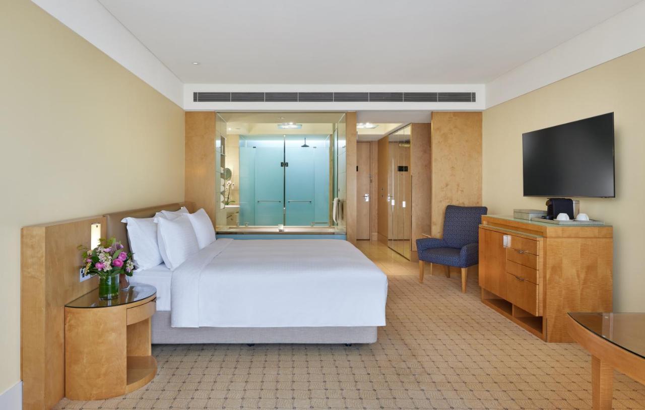 The Fullerton Hotel Sydney Esterno foto The photo shows a modern hotel room. It features a large bed with white linens, a small bedside table with a vase of flowers, and a comfortable blue armchair in one corner. There is a wooden dresser and a flat-screen TV mounted on the wall. Behind th