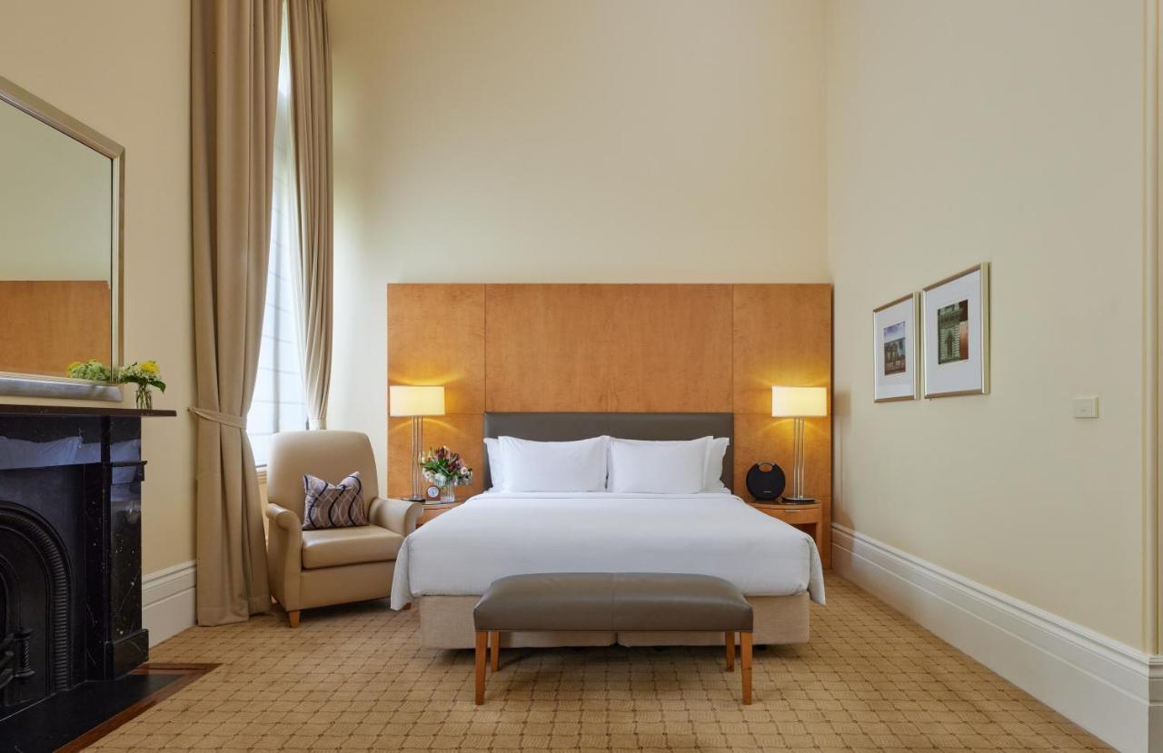 The Fullerton Hotel Sydney Esterno foto The photo shows a well-appointed hotel room. It features a large bed with white linens and a gray headboard, positioned against a wooden wall panel. On either side of the bed are stylish lamps. There is a comfortable armchair in the corner, accompani