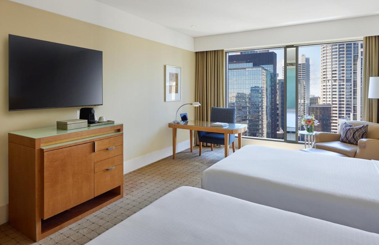 The Fullerton Hotel Sydney Esterno foto The photo shows a hotel room featuring two beds with white bedding. There is a wooden furniture piece with a television mounted on the wall above it. To the side, there is a desk with a chair and a lamp, positioned near a large window that offers a v