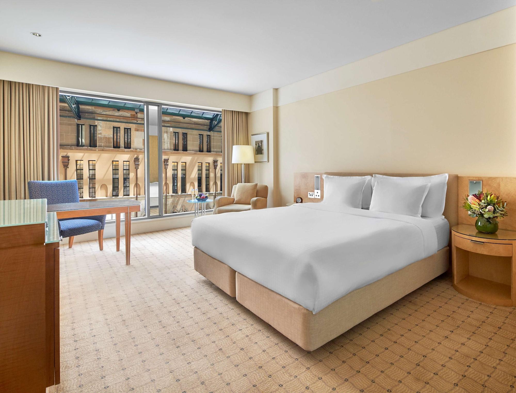 The Fullerton Hotel Sydney Esterno foto The photo shows a hotel room featuring a large bed with white bedding at the center. On one side of the room, there is a light-colored bedside table with a lamp. Across from the bed is a wooden table accompanied by a blue chair, while a comfortable a