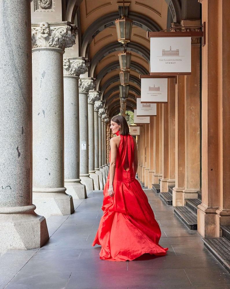The Fullerton Hotel Sydney Esterno foto The photo depicts a woman wearing an elegant red gown, standing in a covered archway lined with columns. She is turned slightly to look back over her shoulder, showcasing the flow and design of the dress. The environment features multiple arches and 