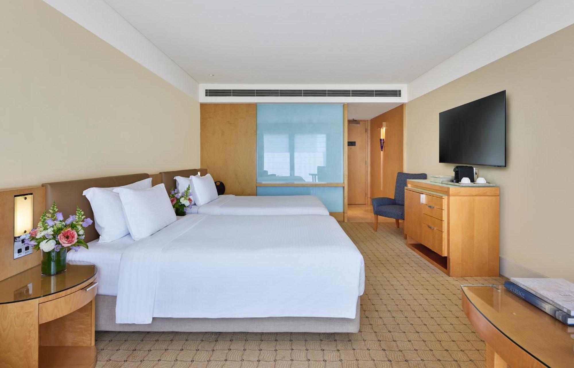 The Fullerton Hotel Sydney Esterno foto The photo shows a hotel room with two beds, each made up with white linens and pillows. There are small flower arrangements on the side tables next to the beds. The room has a light color scheme with beige walls and wooden furniture. A blue glass par