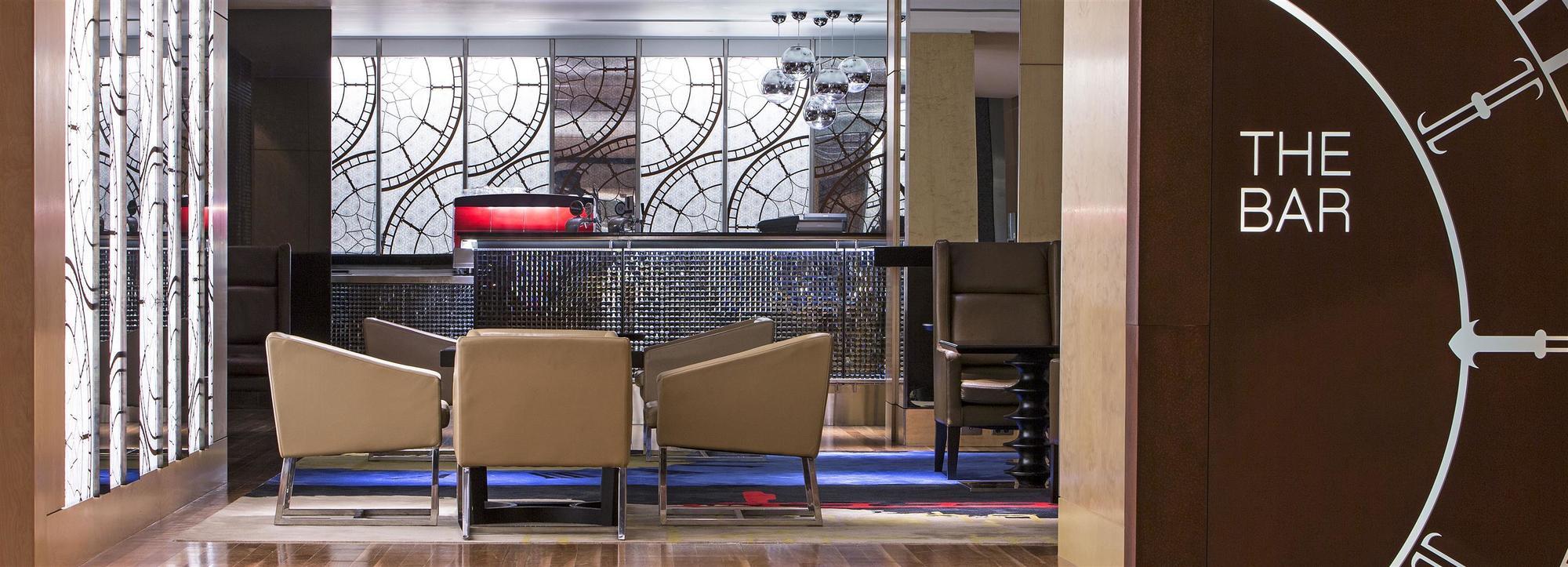 The Fullerton Hotel Sydney Esterno foto The photo shows a stylish interior of a hotel lobby or bar area. In the foreground, there are several beige lounge chairs arranged around a low table, creating a cozy seating area. The background features a modern bar with a sleek, dark counter and a