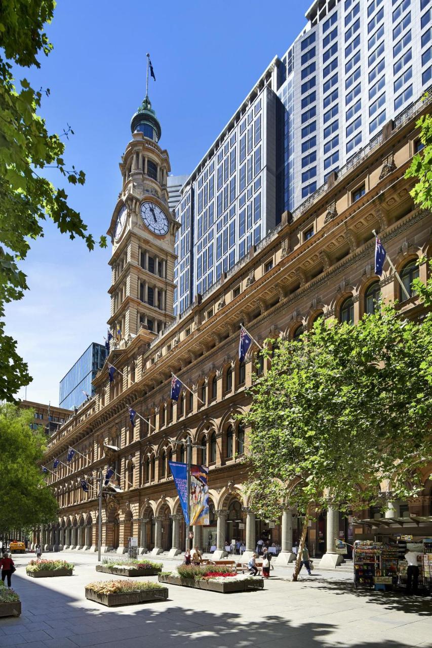 The Fullerton Hotel Sydney Esterno foto The photo shows a historic building featuring a prominent clock tower. The architecture is characterized by ornate details and large windows. The structure has flags on display, and the foreground includes greenery, suggesting that it is set in an ur